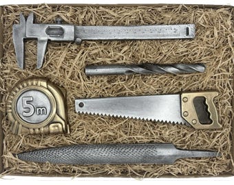 May include: A collection of miniature metal tools, including a caliper, drill bit, tape measure (5m), saw, and file, presented in a cardboard box filled with straw. The tools have a silver finish with brass accents.