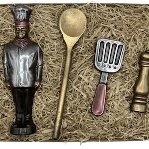 Peut inclure: Une boîte-cadeau remplie de papier de soie brun contient une figurine de chef en chocolat, une cuillère en bois de couleur dorée, une spatule argentée avec un manche rose et un poivrier doré.