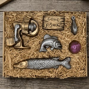 Peut inclure: Un coffret cadeau sur le th&egrave;me de la p&ecirc;che rempli de paille. L'ensemble comprend un moulinet, des ornements en forme de poisson, un poids, une pancarte avec l'inscription "GONE FISHIN" et une boule violette. Les articles sont en m&eacute;tal.