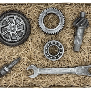 May include: A collection of silver-colored automotive parts, including a wheel, gears, a spark plug, a bearing, and a wrench, arranged in a cardboard box filled with straw-like packing material. The parts have a metallic finish and are detailed.