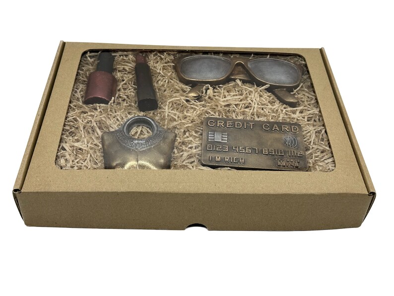 May include: A brown cardboard box with a clear window showing a set of novelty items including a pair of sunglasses, a bottle of nail polish, a lipstick, a necklace, and a credit card with the text "CREDIT CARD" and "I'M RICH".