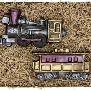 Könnte beinhalten: Ein Set aus zwei Schokoladen-Zügen in einer Schachtel. Die Lokomotive ist silber und schwarz mit einem violetten Schornstein und goldenen Akzenten. Der Personenwagen ist goldfarben mit violetten Akzenten. Beide Wagen haben schwarze Räder.