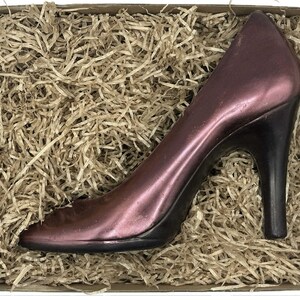 Könnte beinhalten: Ein burgunderfarbener High-Heel-Schuh mit schwarzem Absatz, präsentiert in einem Karton, der mit geschreddertem Papier gefüllt ist. Der Stiletto hat eine spitze Zehenpartie und ein Metallic-Finish.