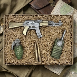 Könnte beinhalten: Eine Sammlung militärischer Gegenstände, präsentiert in einer Box mit Stroh. Das Set umfasst ein silber- und goldfarbenes Gewehr, eine grüne Granate, zwei Patronen und ein grünes Walkie-Talkie. Die Gegenstände sind auf einem Strohbett angeordnet.