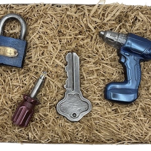 Op de afbeelding: Een cadeaudoos gevuld met bruin verpakkingsmateriaal en vier gereedschappen: een blauw-zilveren boormachine, een zilveren sleutel, een rode schroevendraaier en een blauw hangslot met het woord "BULL" erop.