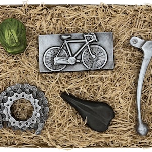 May include: A gift box filled with chocolate bicycle themed gifts. The box contains a chocolate bicycle, a chocolate chain, a chocolate helmet, a chocolate seat, and a chocolate brake lever. All the chocolate pieces are silver in color.