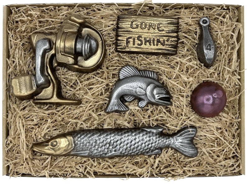 Peut inclure: Une collection d'objets sur le th&egrave;me de la p&ecirc;che dans une bo&icirc;te en carton remplie de paille. Les objets comprennent un moulinet, une pancarte avec l'inscription "Gone Fishin'", un poids marqu&eacute; "4", un poisson, une boule et un long poisson.