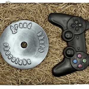 Könnte beinhalten: Ein brauner Karton mit zerknülltem Papier im Inneren. Die Schachtel enthält eine silberne Scheibe mit den Worten "Grand Theft Chocolate" und einen schwarzen Schokoladen-Videospielcontroller.