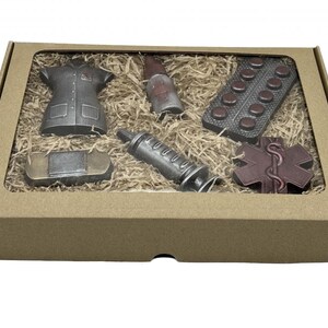 Puede incluir: Una caja de cart&oacute;n marr&oacute;n con una ventana transparente que muestra un conjunto de figuritas de chocolate. Las figuritas incluyen un uniforme de enfermera, una jeringa, una botella de medicina, una venda, un paquete de pastillas y un s&iacute;mbolo m&eacute;dico rojo.