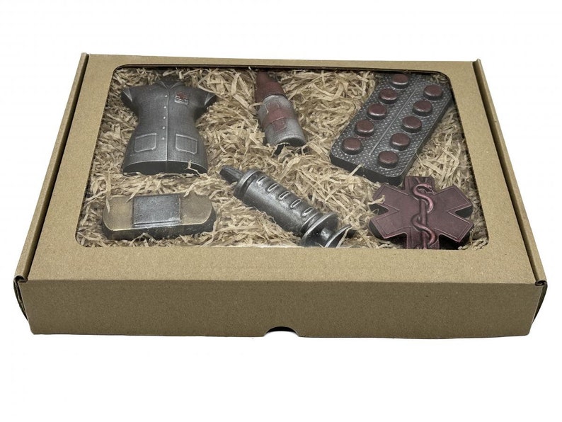 Puede incluir: Una caja de cart&oacute;n marr&oacute;n con una ventana transparente que muestra un conjunto de figuritas de chocolate. Las figuritas incluyen un uniforme de enfermera, una jeringa, una botella de medicina, una venda, un paquete de pastillas y un s&iacute;mbolo m&eacute;dico rojo.