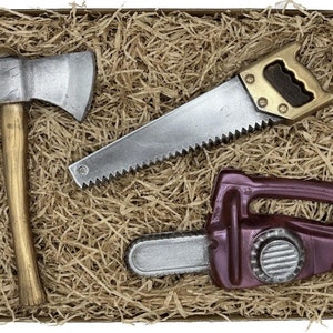 Peut inclure: Un ensemble de trois outils en chocolat dans une bo&icirc;te brune avec du papier d&eacute;chiquet&eacute;. Les outils sont une hache argent&eacute;e, une scie &agrave; main argent&eacute;e et une tron&ccedil;onneuse violette.