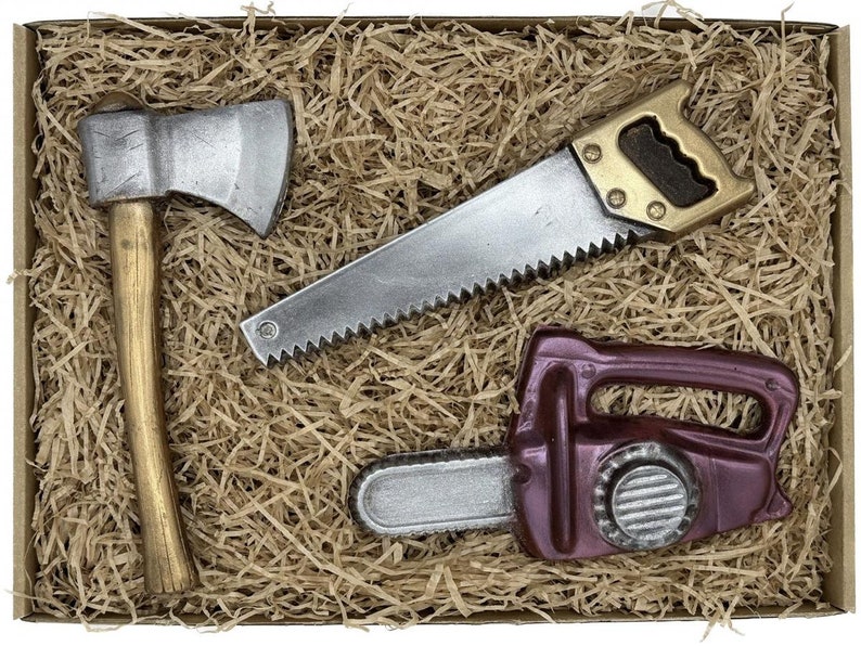 Peut inclure: Un ensemble de trois outils en chocolat dans une bo&icirc;te brune avec du papier d&eacute;chiquet&eacute;. Les outils sont une hache argent&eacute;e, une scie &agrave; main argent&eacute;e et une tron&ccedil;onneuse violette.