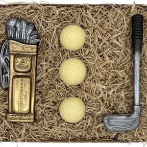 Könnte beinhalten: Eine Geschenkbox gefüllt mit braunem Seidenpapier, die einen goldenen Golfsack, drei weiße Golfbälle und einen silbernen Golfschläger enthält.