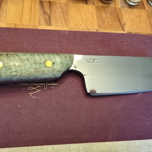 May include: A stainless steel cleaver knife with a gray and green wooden handle, secured with two gold-colored rivets. The blade has a rectangular shape and the text "507" is etched on the blade. The knife rests on a burgundy surface.
