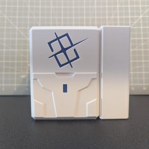 Op de afbeelding: Een wit rechthoekig plastic object met een donkerblauw geometrisch symbool op de voorzijde. Het object heeft gegoten paneellijnen en een klein donkerblauw rechthoekig accent nabij het midden onderaan, naast een bijpassend wit zijstuk.