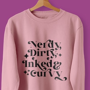 May include: A pink sweatshirt with the text "Nerdy, Dirty, Inked & Curvy" printed in black on the front. The sweatshirt is hanging on a hanger. The text "Create cute t-shirts" is printed below the sweatshirt.