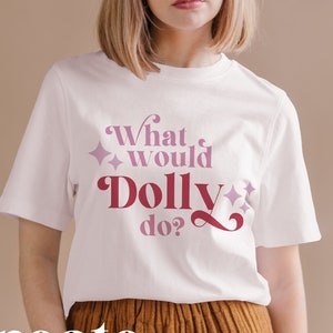 Pode incluir: Camiseta branca com o texto "What would Dolly do?" em letras rosa e vermelhas com detalhes de glitter.