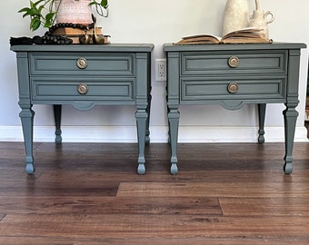 May include: Two matching teal bedside tables with two drawers each and brass knobs. The tables have ornate legs and are topped with decorative items, including a potted plant, books, and a vase with branches.