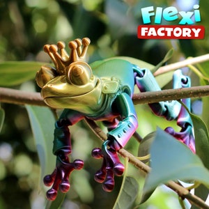 May include: A green, gold, and purple 3D printed frog wearing a gold crown. The frog is perched on a branch with green leaves. The text "Flexi FACTORY" is in the background.