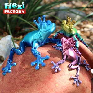 May include: Three articulated frog figurines with crowns, each painted in a different iridescent color: blue, green, and purple. The frogs are posed on a red-brown surface. The text "Flexi Factory" is visible in the background.