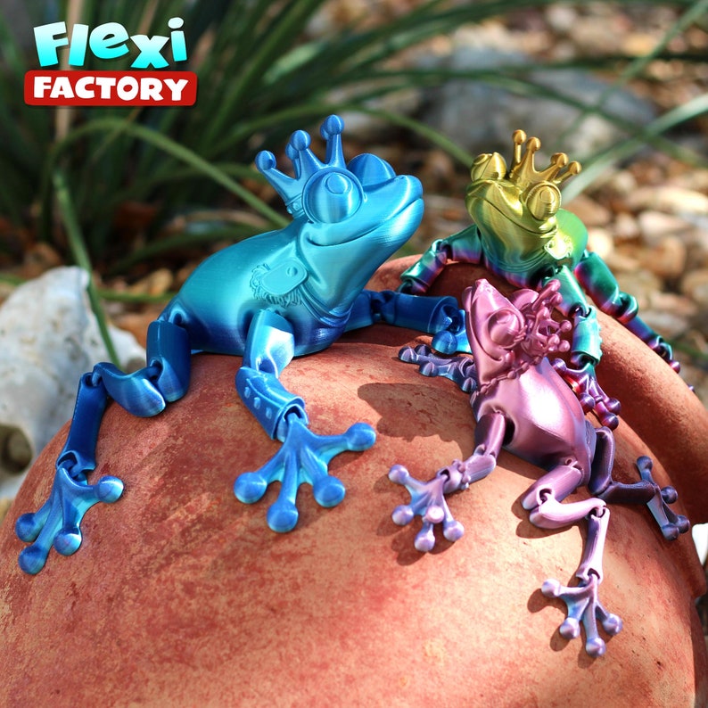 May include: Three articulated frog figurines with crowns, each painted in a different iridescent color: blue, green, and purple. The frogs are posed on a red-brown surface. The text "Flexi Factory" is visible in the background.