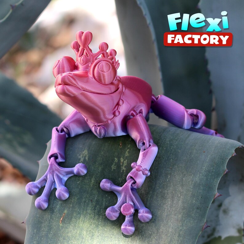May include: A pink and purple articulated frog figurine wearing a crown and necklace. The frog is sitting on a green leaf. "Flexi Factory" is written in red letters on a white background in the upper right corner of the image.