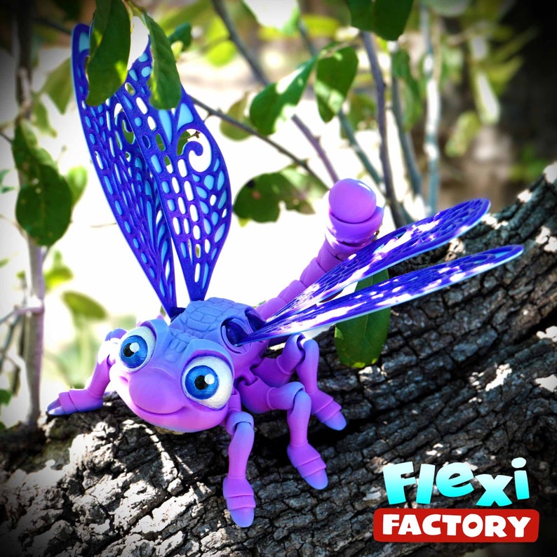 May include: A purple and blue dragonfly toy with large eyes and articulated legs and wings. The dragonfly is perched on a tree branch. The wings are a vibrant blue with a detailed, openwork design. The logo "Flexi Factory" is visible.