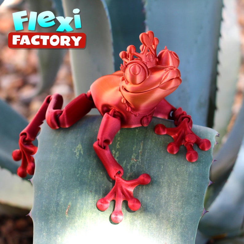 May include: A red articulated frog figurine wearing a crown and necklace, sitting on a green leaf. The figurine is made by Flexi Factory.