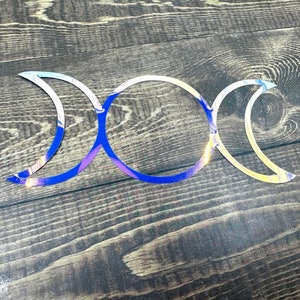 May include: Triple moon symbol made of iridescent vinyl with a blue, purple, and yellow color scheme. The symbol is on a dark wood background.
