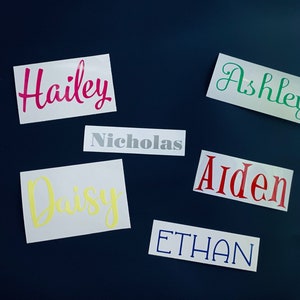 May include: Six white labels with different names written in various colors. The names are Hailey in pink, Ashley in green, Nicholas in gray, Daisy in yellow, Aiden in red, and Ethan in blue.