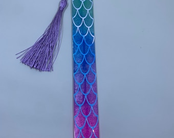May include: A mermaid-themed bookmark with a gradient of blue, green, and purple colors. The bookmark has a tassel attached to the top.