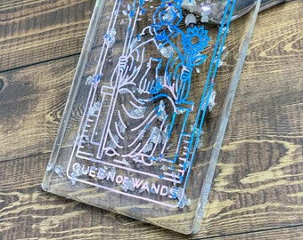 May include: A clear resin coaster with a Queen of Wands tarot card design. The card is outlined in blue and pink and has a silver glitter background.