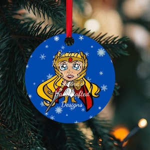 May include: A round blue Christmas ornament featuring a cartoon character with long blonde hair, a gold headpiece, and a red cape on one side, and the text "Mini Masters of the Universe Designs", "NATALIE", and "2023" on the reverse side.