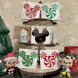 May include: Four wooden squares with a Christmas-themed Mickey Mouse design. Each square features a different design, including a candy cane Mickey Mouse, a black Mickey Mouse with a red nose, and two candy cane Mickey Mouse designs with different color schemes. The squares are displayed on a tiered wooden stand.