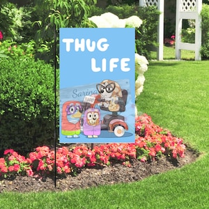 May include: A garden flag with the words "THUG LIFE" in white on a light blue background. The flag features cartoon characters, including one in a motorized scooter. The flag is displayed in a garden with flowers and green grass.