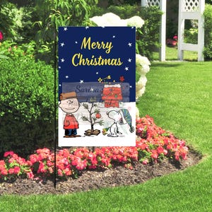 May include: A garden flag with a navy blue background and the words "Merry Christmas" in yellow. The flag features a cartoon of Charlie Brown, Snoopy, and a Christmas tree. The flag is displayed in a garden with flowers.