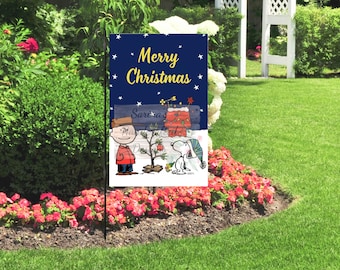 May include: A garden flag with a navy blue background and the words "Merry Christmas" in yellow. The flag features a cartoon of Charlie Brown, Snoopy, and a Christmas tree. The flag is displayed in a garden with flowers.