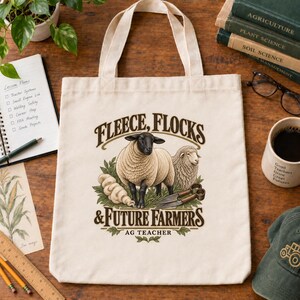 May include: A cream-colored canvas tote bag featuring a graphic of two sheep, a pair of metal shears, and green foliage. The text reads: Fleece, Flocks & Future Farmers, Ag Teacher. The design is centered on the front of the bag with two handles.