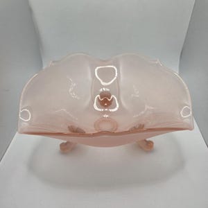 May include: A light pink, footed glass bowl with a scalloped rim. The bowl has a decorative design in the center and is supported by three small feet. The glass has a translucent quality.