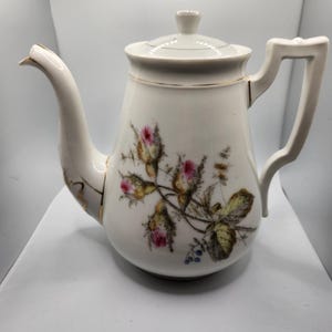 May include: White porcelain teapot with a floral design featuring pink roses and green leaves. The teapot has a gold trim around the spout and handle.