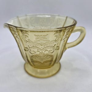 May include: A vintage yellow glass creamer with an ornate embossed floral design. The creamer has a handle and a spout for easy pouring. The base is a pedestal shape. The glass has a slight amber tint.