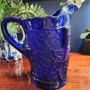 May include: A cobalt blue glass pitcher with a textured, honeycomb-like design and a floral motif. The pitcher has a wide mouth and a handle.