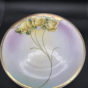 May include: A round, decorative plate with a gold rim and a floral design. The plate features three yellow roses with green leaves and stems. The background has a gradient of white, lavender, and light blue.