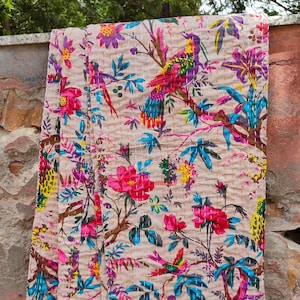 Könnte beinhalten: Eine beige Kantha-Steppdecke mit einem floralen und Vogelmuster. Die Steppdecke ist in den Farben Pink, Blau, Gelb und Grün gehalten. Die Steppdecke ist gefaltet und hängt an einer Steinwand.