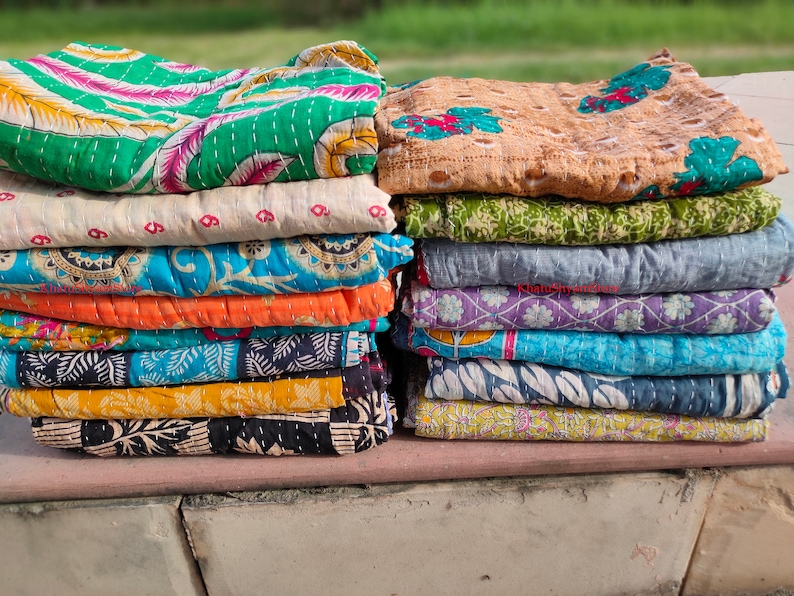 May include: Stacks of colorful, patterned quilts. The quilts feature various designs, including floral and geometric patterns, in shades of green, blue, orange, and yellow. Each quilt is folded and stacked neatly, showcasing the different colors and designs.