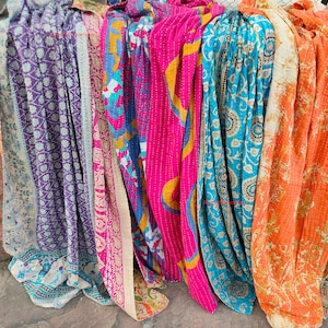 May include: A collection of colorful, patterned textiles, likely blankets or throws, in shades of purple, pink, blue, and orange. Each piece features unique designs and stitching, suggesting a handcrafted or vintage aesthetic. The textiles are draped, showcasing their textures and patterns.
