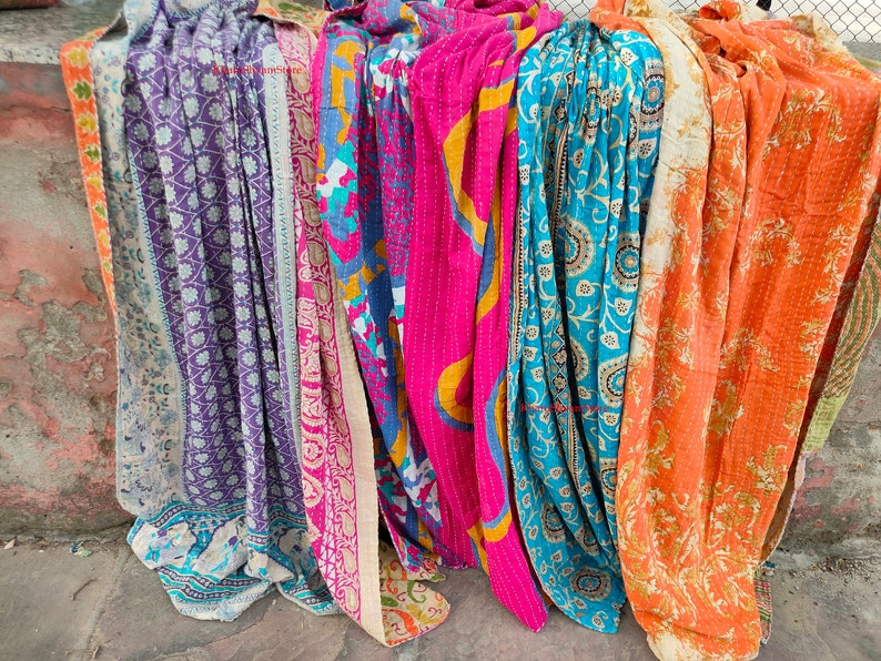 May include: A collection of colorful, patterned textiles, likely blankets or throws, in shades of purple, pink, blue, and orange. Each piece features unique designs and stitching, suggesting a handcrafted or vintage aesthetic. The textiles are draped, showcasing their textures and patterns.