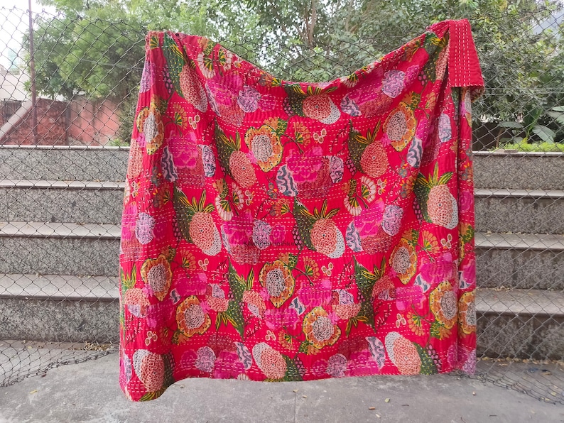 Op de afbeelding: Een rode kantha-deken met een bloemenpatroon met roze, gele en groene bloemen. De deken is gemaakt van katoen en met de hand gestikt.