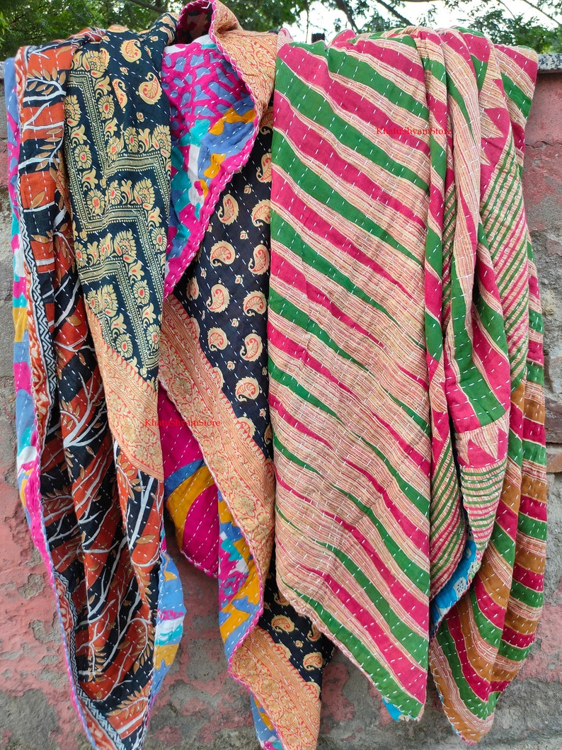 May include: A collection of colorful, patterned quilts. The quilts feature various designs, including paisley, floral, and striped patterns. The color palette includes shades of blue, pink, green, and gold. The quilts are hanging, showcasing their textures and designs.