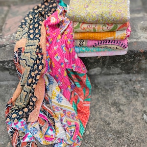 May include: A stack of colorful, patterned quilts and a folded quilt. The quilts feature various designs, including floral and geometric patterns, in shades of pink, yellow, orange, and black. The quilts are made of soft fabric.